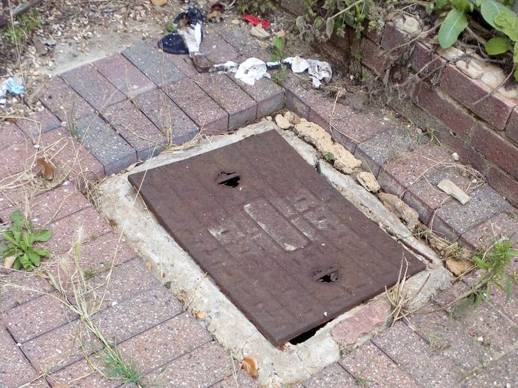 A rusted inspection chamber cover set into brick paving beside a garden bed